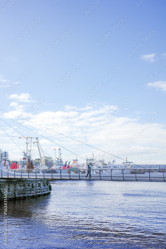 Fototapeta premium Scenic harbor view at the VA Waterfront in Cape Town, South Africa, featuring a modern pedestrian swing bridge, docked ships, and bright blue skies. The calm water reflects the maritime atmosphere