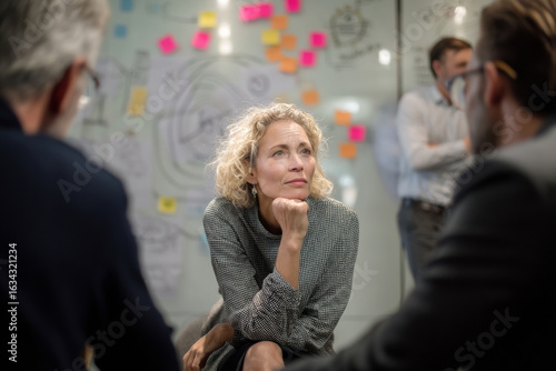 Middle-aged Caucasian businesswoman seated in a circular team huddle, participating in a project retrospective while offering thoughtful reflections, with sticky notes and whiteboard sketches all