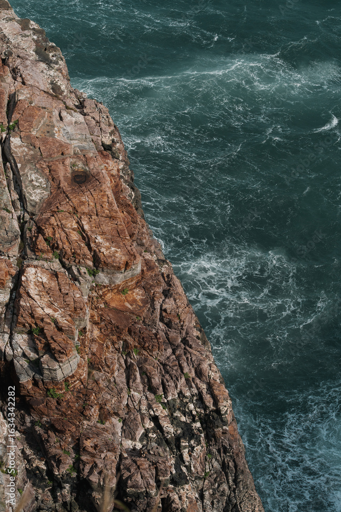 Tableau sur toile Dramatic coastal view of a rugged, reddish-brown cliff face meeting turbulent, dark ocean waves