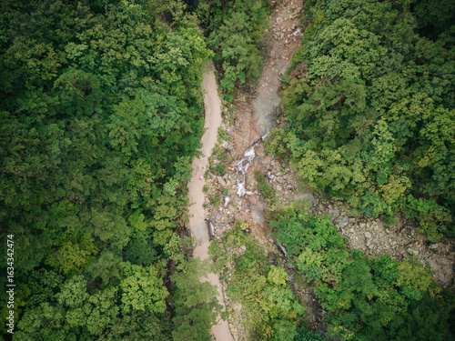 Aerial view of a dirt path winding through a lush, forested ravine with a small waterfall.