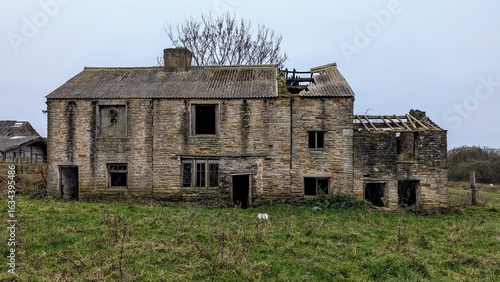 Abandoned collapsing country farmhouse, Lancashire UK. Derelict rural farm cottage building in grass field. Falling down countryside house. Uninhabited burnt out stone shell ruin, wrecked roof decay.