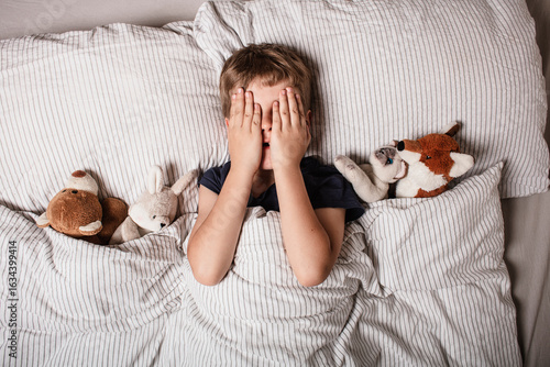 The boy was scared before going to bed. Night terrors in a child. The kid covers his face with his hands in a fear. Children's experiences. Boy in the bed. View from above. Sad psychological state.