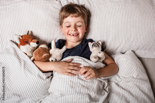 Happy child waking up in bed with big smile and raised arm, surrounded by stuffed animals. Joyful morning energy, childhood happiness and cozy home atmosphere. Fun before a sleeping. Kid smile in bed.