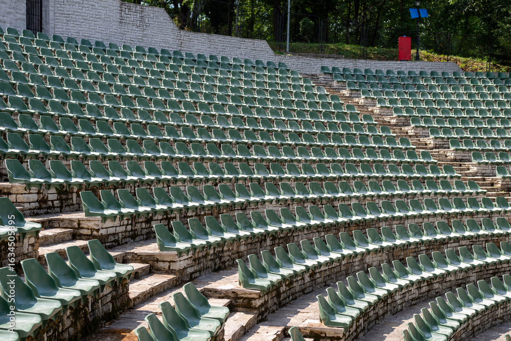 Fototapeta premium Large, open-air stone amphitheater is filled with numerous rows of green plastic seats arranged in a semi-circular pattern