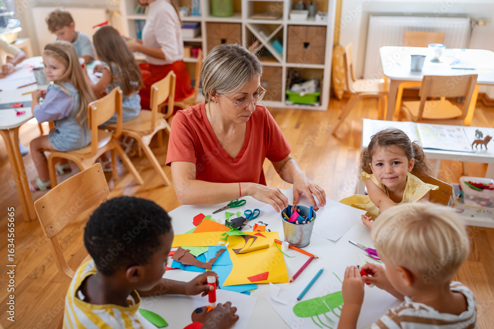 Fototapeta premium Preschool teacher engaging with children at a table during an arts and crafts activity in a bright classroom