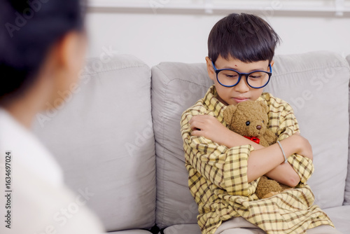 Fotografía Asian boy with ADHD sitting alone on sofa hugging teddy bear displaying sadness