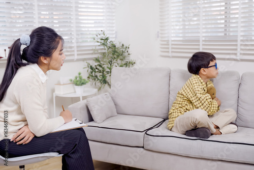 Asian female psychologist sitting beside young Asian boy with ADHD during therapy session on sofa in clinic, writing notes while observing withdrawn child hugging stuffed toy silently