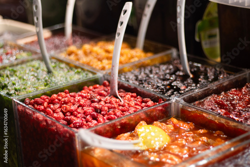 Cuadro en lienzo Assorted preserved fruits and syrups presented in square acrylic bins at a market counter, including bright red berries, orange citrus peel, and green chopped zest