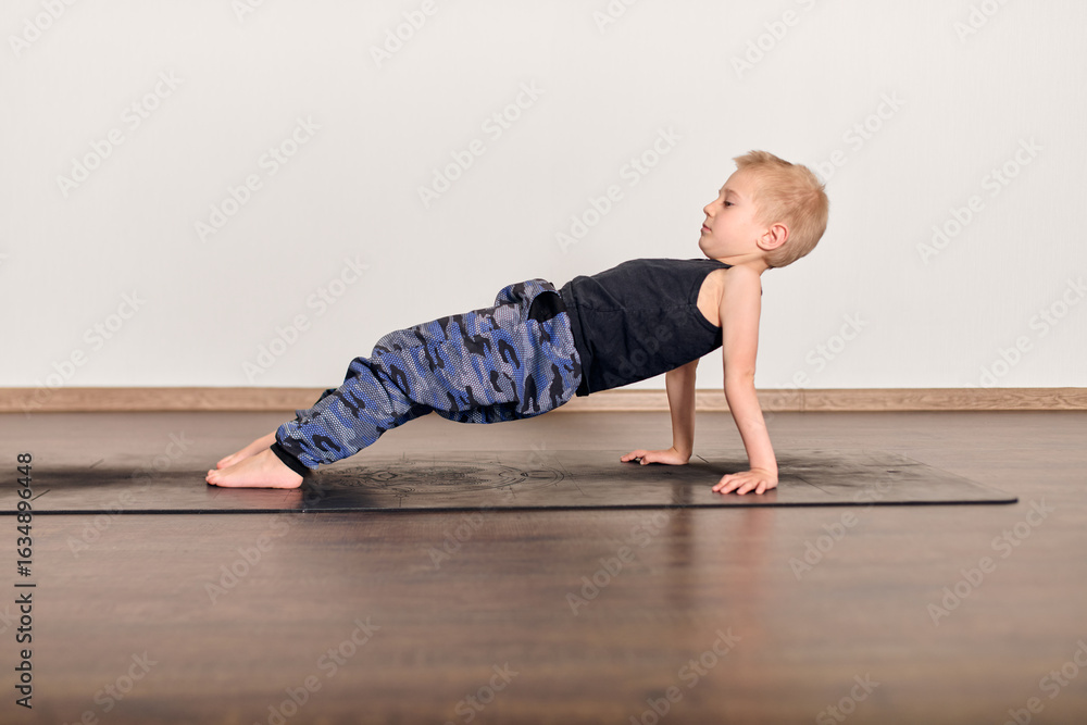 Naklejka premium Child with blonde hair performs yoga on mat, wearing casual outfit. Indoor studio with wooden floor and light walls creates serene atmosphere. Focus on balance and concentration