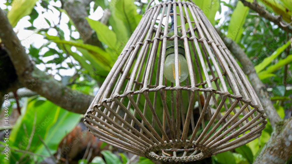Obraz premium Close up of a woven rattan pendant lights with green plant bacground. A lampshade woven from rattan. A hanging lamp for outdoor use, home gardens, restaurants and decoration of a room's. interior. 