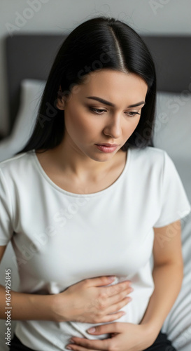 Free Download Close-up of a young woman with dark hair, wearing a white shirt, holding her stomach, conveying discomfort or pain.  Represents health concerns, wellness, or medical issues. - GFXLoad