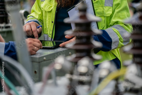 Utility workers in high visibility safety jackets using tablet device for inspection, diagnostics, and maintenance planning at outdoor electrical substation power distribution equipment