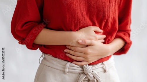 Woman in red shirt holding her stomach with a concerned expression, indicating discomfort or pain in a softly lit indoor space