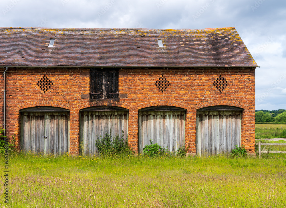 Obraz premium A weathered brick barn with four wooden doors sits in a field in the English countryside of Shropshire