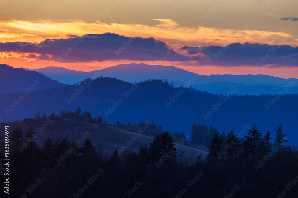 Obraz premium Layered mountain ranges illuminated by vibrant sunrise, casting long shadows in the twilight scenery.