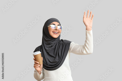 Beautiful Muslim woman in eyeglasses with cup of coffee on grey background