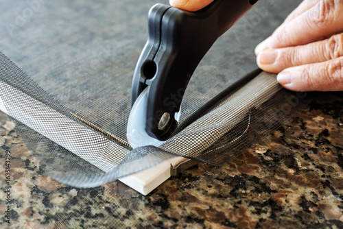 Close up view as a worker rescreens and replaces the mesh of a window screen with a spline roller tool on a white window frame.
