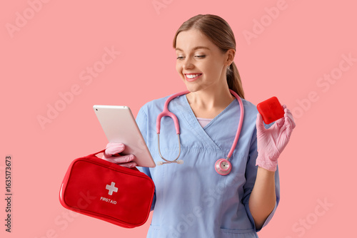 Female doctor with first aid kit, pillbox and tablet computer on pink background