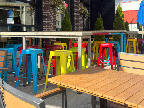 Bright, colorful, iron metal stackable bar stools; yellow, blue, and red colored. The chairs are around a tall bar table on a restaurant patio. There's garden furniture made of light wood and metal. 