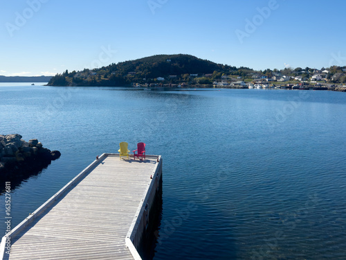 A sturdy wooden pier juts out into the still water of a lake. The wharf has both yellow and red Adirondack chairs along its edge. The bright blue water is a sheltered cove with tree-covered hills.