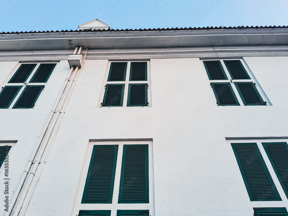 Fototapeta premium Fatahillah Museum or Jakarta History Museum facade in Kota Tua with colonial architecture, green wooden shutters, and white walls under blue sky