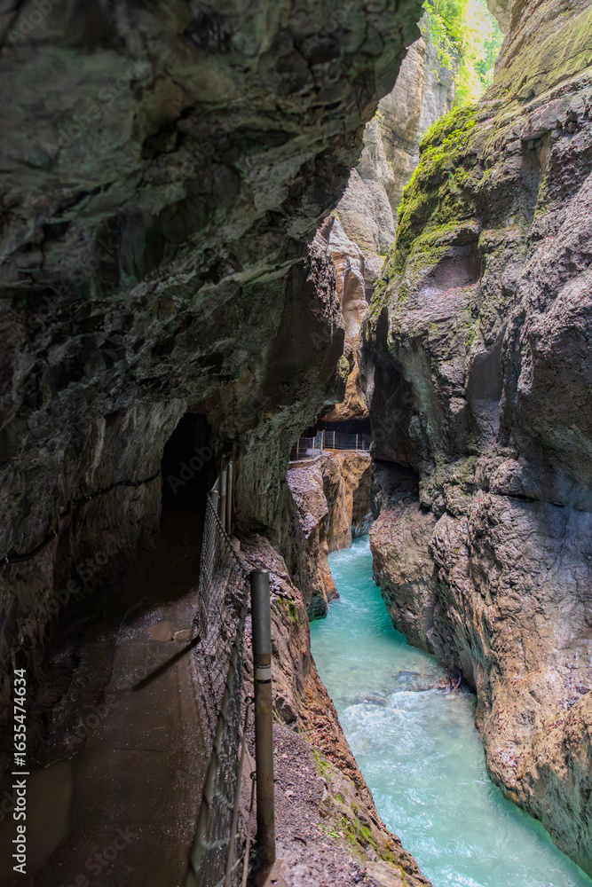 Fototapeta premium Narrow walkway carved into the cliffside along a turquoise stream inside Partnachklamm Gorge, Garmisch-Partenkirchen, Bavaria. A stunning natural passageway through dramatic alpine rock formations.