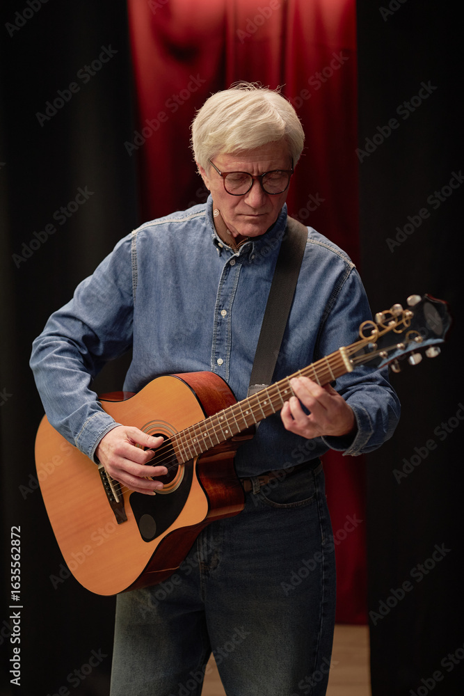 Obraz premium Mature Caucasian man playing acoustic guitar on stage during talent competition, wearing glasses and focusing on performance, standing against dark background with red curtain