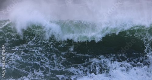 An large ocean storm wave curls and breaks over a coastal rock ledge, taking the appearance of a huge waterfall, before exploding skyward into a giant wall of white water. Captured in slow motion.
