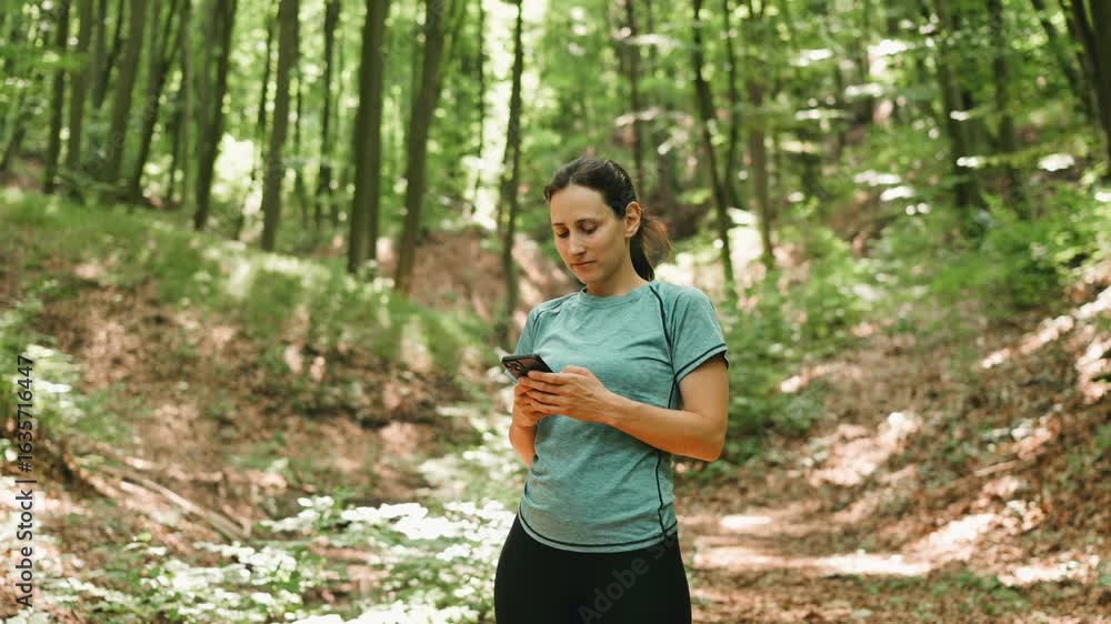 A lively young woman dressed in trendy activewear is checking her smartphone in a peaceful forest setting A lively young woman dressed in trendy activewear is checking her smartphone in a peaceful forest setting
