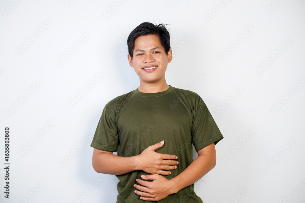 Obraz premium A cheerful young Asian man wearing a green shirt smiles while holding his stomach, standing against a plain white background, exuding happiness and good health. 