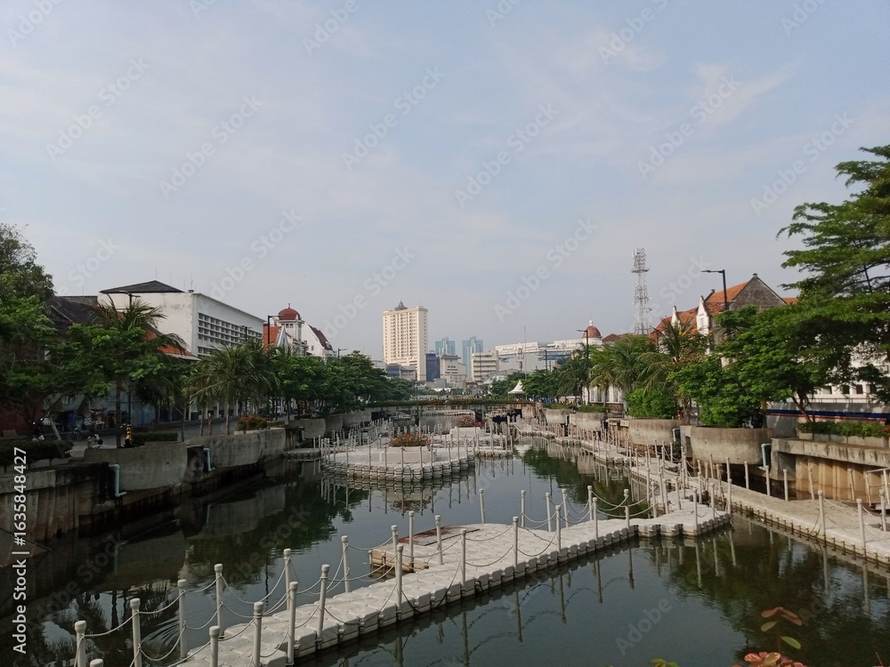 Obraz premium A panoramic shot of the Kali Besar river in Jakarta's Old Town, with the water flanked by walkways, floating platforms, and historical Dutch colonial-style buildings under a hazy sky
