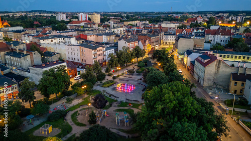 Aerial view of Ostrów Wielkopolski in the evening, featuring a park with a colorful fountain and playground, surrounded by urban buildings.