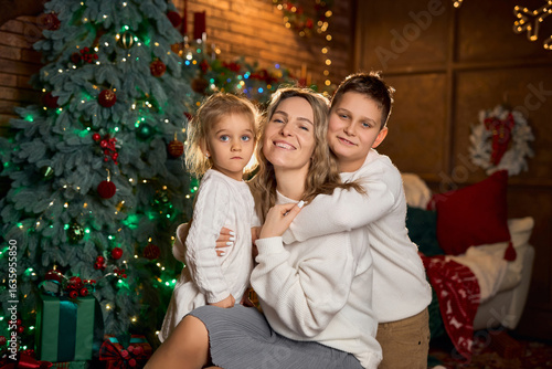 smiling mother and her two children in white sweaters hug closely, celebrating in a cozy, festive room with a decorated tree and golden lights
