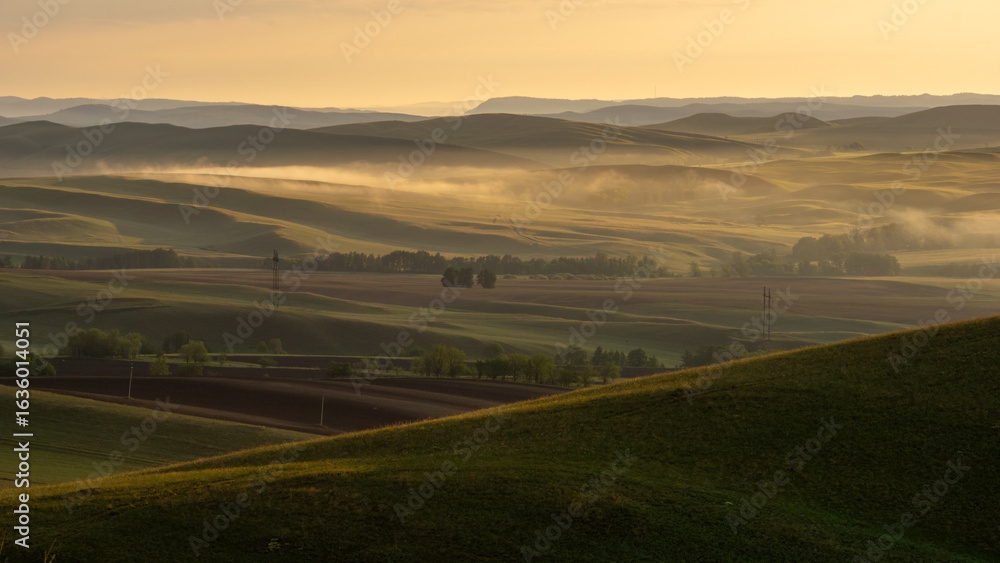Fototapeta premium Long mountains. Serene landscape at dawn, showcasing rolling hills covered in lush greenery, with soft mist rising above fields, creating a tranquil and picturesque rural scene
