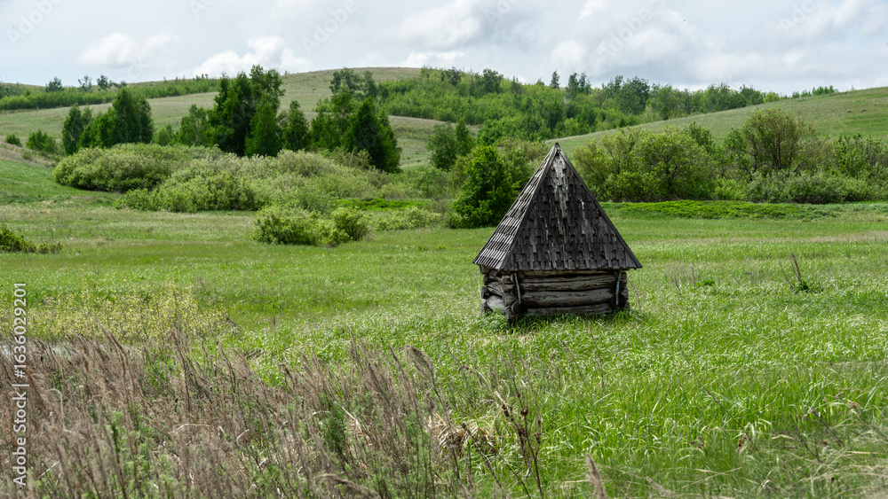 Obraz premium Rustic wooden structure with a triangular roof stands alone in a lush green meadow, surrounded by rolling hills and trees under a bright cloudy sky, evoking tranquility and solitude