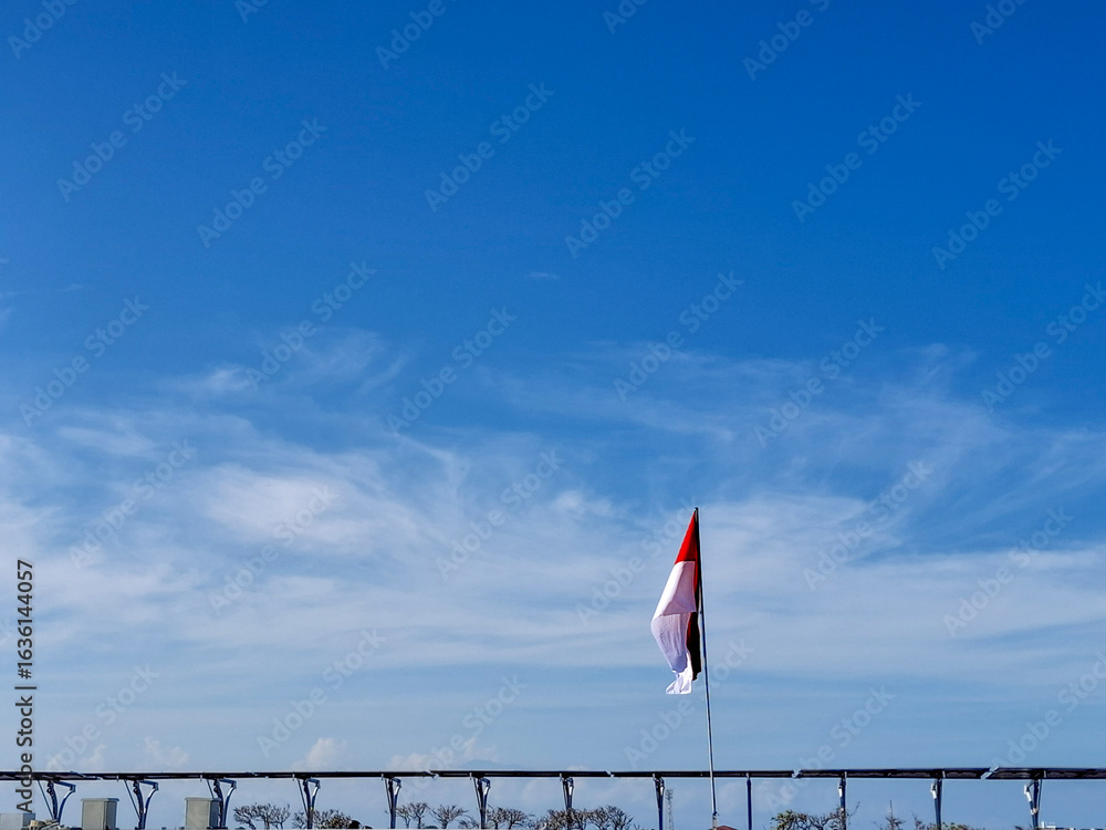 Fototapeta premium A lone red and white flag stands proudly against a vast, clear blue sky with soft, wispy clouds. The flag, a symbol of freedom and resilience, flutters gently in the wind