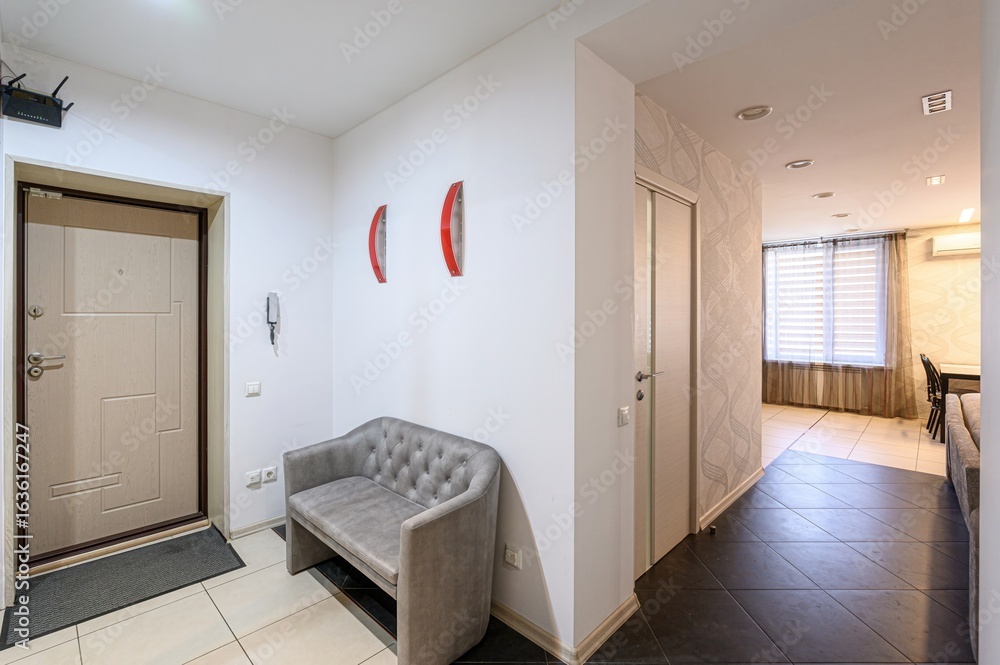 interior shot reveals an apartment entry with a white door, grey bench, and a glimpse into the living area. The decor is minimal, highlighting the tiled floors and neutral tones