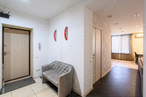 interior shot reveals an apartment entry with a white door, grey bench, and a glimpse into the living area. The decor is minimal, highlighting the tiled floors and neutral tones