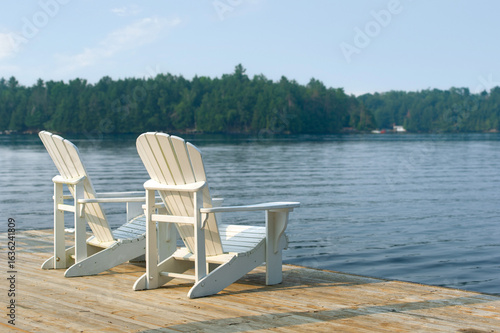 White Adirondack chairs perched on a dock overlook a forest-lined lake in Ontario, basking in the warm glow of early morning sun. The scene captures the essence of a serene summer retreat.