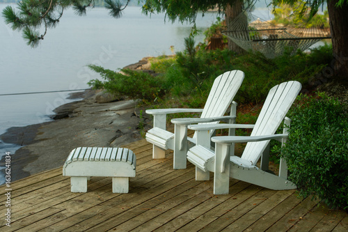 Classic Muskoka chairs rest under tall pines on a sun-dappled wooden deck, facing the gentle ripples of a Kawarthas lake — a perfect cottage country escape.