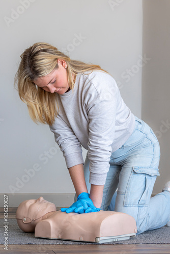 Full-length portrait of a female first aid instructor demonstrating correct posture and hand position for administering chest compressions on a Basic Life Support (BLS) CPR manikin. 