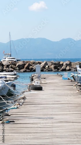 Wallpaper Mural Wooden pier in Aegina Island harbor with moored boats, stone breakwater, calm blue sea, and hazy mountains under a clear sky with a small white cloud, no people. Vertical Torontodigital.ca