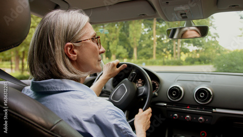 Back view Caucasian mature elderly business woman aged female businesswoman in glasses taxi driver hold steering wheel steer auto inside automobile rearview mirror carefully driving machine attention