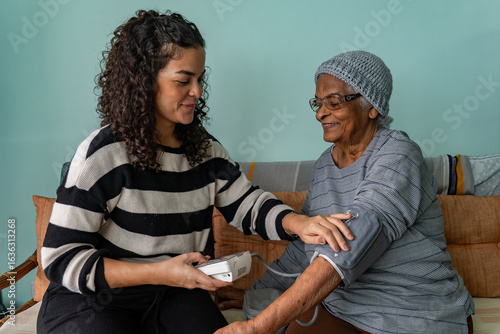Foto Candid Moment of Latina Woman Checking Blood Pressure of Afro Grandmother at Hom