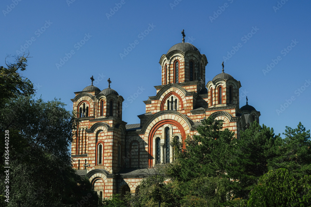 Fototapeta premium St. Mark’s Church in Belgrade, Serbia, with its characteristic red and cream-colored Byzantine-style architecture, stands tall against a clear blue sky, framed by green trees
