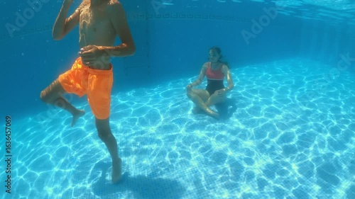 Young boy in orange shorts dancing playfully underwater beside teenage girl meditating in one-piece swimsuit at swimming pool bottom