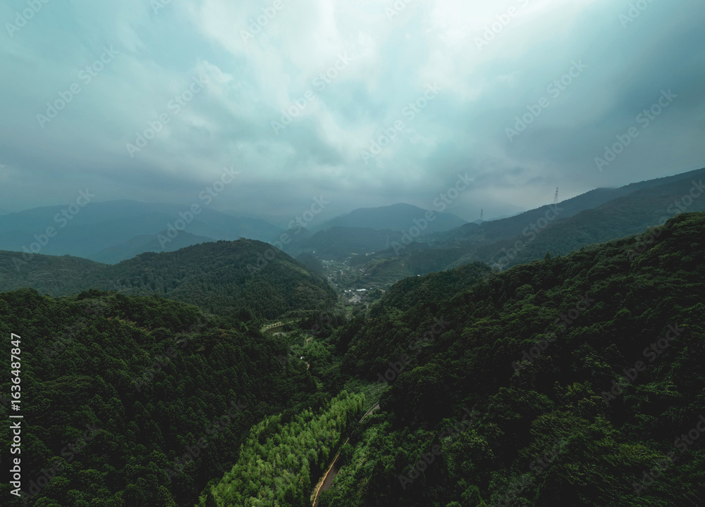 Naklejka premium A vast, misty mountain range shrouded in clouds, with dense green forests and a winding road. A small village nestled in the valley below. 