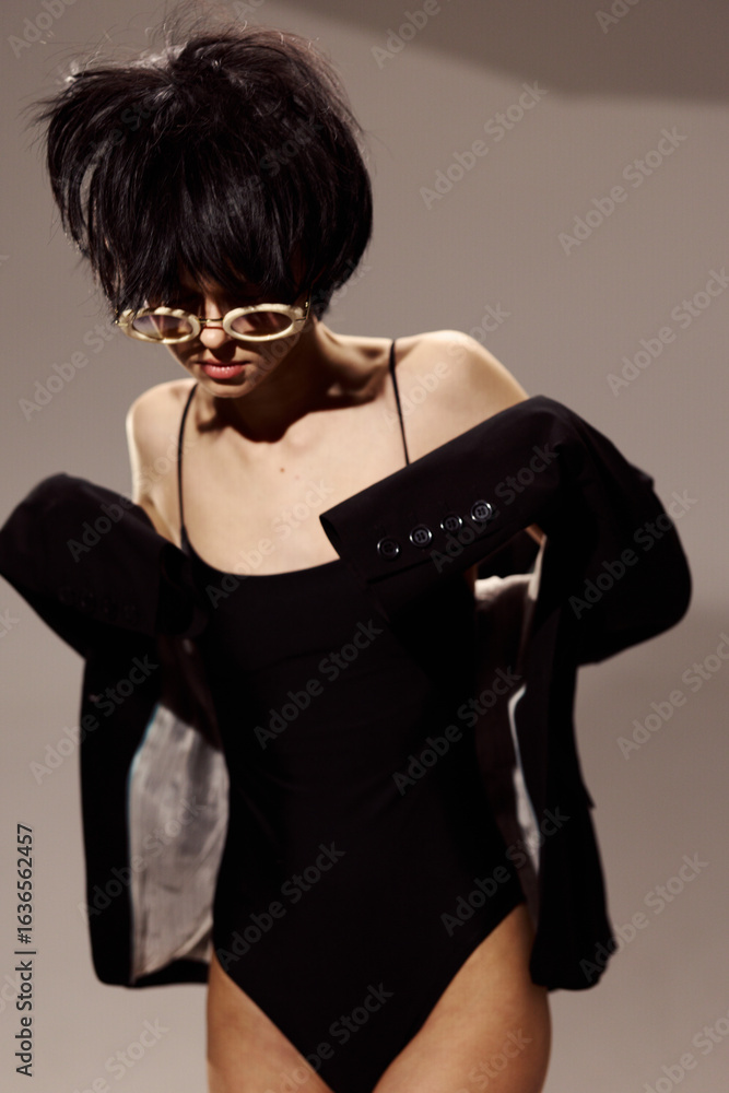 Stylish woman in black bodysuit and jacket posing with short hair and sunglasses on gray background. Stylish woman in black bodysuit and jacket posing with short hair and sunglasses on gray background.