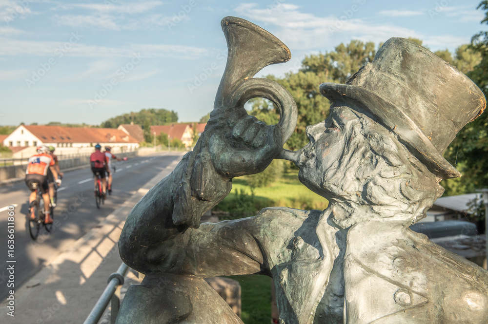 Obraz premium Skulptur eines Postillons mit Zylinder und Uniform der in ein Posthorn bläst. Im Hintergrund eine Sandsteinbrücke mit unkenntlichen Radfahrern vor blauem Himmel mit Schleierwolken in Neckartailfingen.