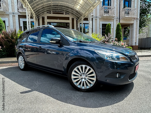 A stylish blue hatchback is elegantly parked right in front of a luxurious and grand building, surrounded by beautifully maintained landscaping that really enhances the vibrant urban environment